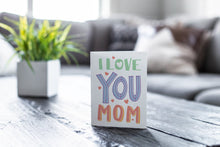 Load image into Gallery viewer, A greeting card featured on a black, wood coffee table. There’s a white planter in the background with a green plant. There’s also a gray sofa in the background with a white pillow. The card features the words “I love you mom.” 