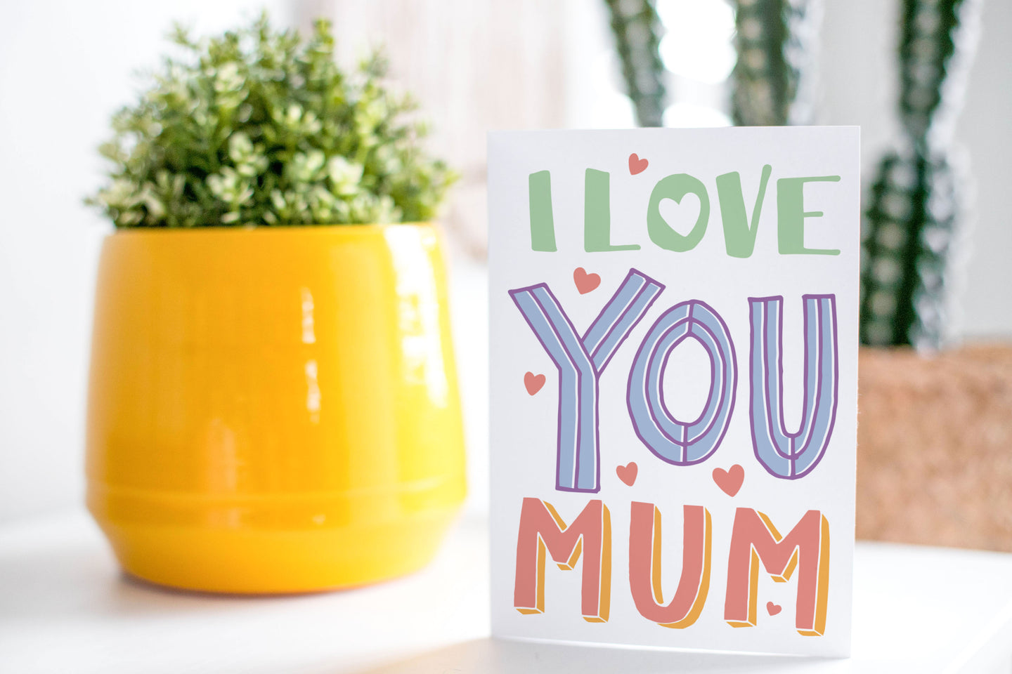 A greeting card is on a table top with a yellow plant pot and a green plant inside. The card features illustrated lettering reading “I love you Mum” with small hearts around the words.