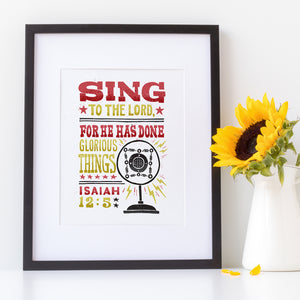 Artwork in a black frame with the with a white matte. The artwork is on a white background with lettering reading "Sing to the Lord for he has done glorious things. Isaiah 12:5." The words are in red, yellow and black. 
