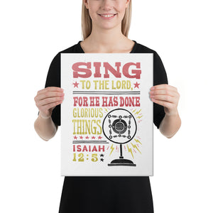 A woman holding a canvas in her hands. The canvas has a white background with the words "Sing to the Lord for he has done glorious things. Isaiah 12:5." The words are in red, yellow and black. 