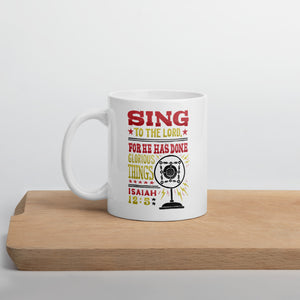 A white mug sitting on a piece of wood. The white mug features hand drawn lettering with the words "Sing to the Lord for he has done glorious things. Isaiah 12:5." The words are in red, yellow and black.