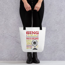 Load image into Gallery viewer, Someone holding a tote bag with black handles and a white fabric bag. The artwork features lettering red, yellow and black reading hand drawn lettering with the words &quot;Sing to the Lord for he has done glorious things. Isaiah 12:5.&quot; 