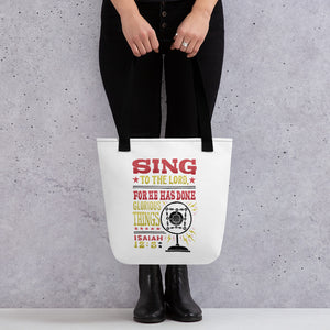 Someone holding a tote bag with black handles and a white fabric bag. The artwork features lettering red, yellow and black reading hand drawn lettering with the words "Sing to the Lord for he has done glorious things. Isaiah 12:5." 
