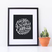 Load image into Gallery viewer, Artwork in a white frame with the with a white matte. The frame is leaning on a white tabletop with a terracotta pot with a cactus. The artwork is on a black background with some white texture to give a vintage look. The text is in white and reads “Live a life worthy of the calling you have received.” 