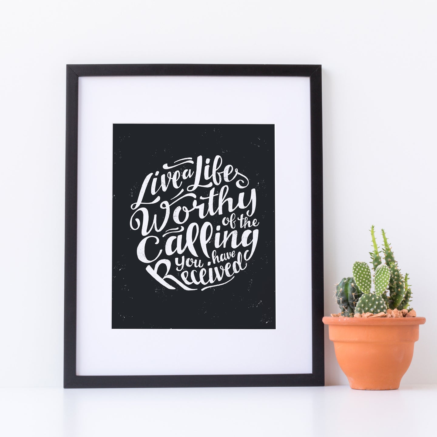 Artwork in a white frame with the with a white matte. The frame is leaning on a white tabletop with a terracotta pot with a cactus. The artwork is on a black background with some white texture to give a vintage look. The text is in white and reads “Live a life worthy of the calling you have received.” 