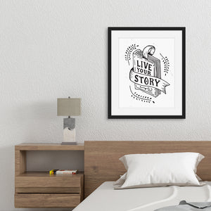 Artwork featured in a black frame in a bedroom above a bed. The artwork is printed on white paper and features black and red hand drawn lettering with the words "Live Your Story" inside an illustrated book. 
