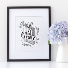 Load image into Gallery viewer, Artwork in a black frame with the with a white matte. The frame is leaning on a shelf with a white pot of light purple flowers next to it. The artwork is on a white background with lettering reading &quot;Live Your Story&quot; The words are inside an illustrated book.