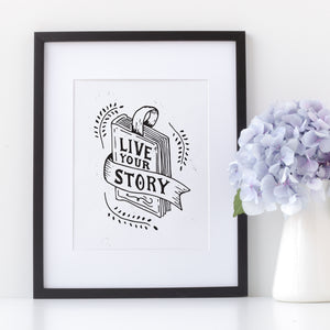 Artwork in a black frame with the with a white matte. The frame is leaning on a shelf with a white pot of light purple flowers next to it. The artwork is on a white background with lettering reading "Live Your Story" The words are inside an illustrated book.