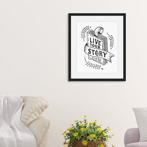 The artwork is featured in a black frame hanging on the wall with the words "Live Your Story" inside an illustrated book. The artwork is hanging above a basket of yellow flowers and two sofa chairs. 