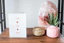 Load image into Gallery viewer, A card on a wood tabletop and on the right side of the card is a woven basket, a pink plant pot with a cactus in it and a pink crystal rock. The card features the words “Love You Mum” with a small, red heart in between each word. 