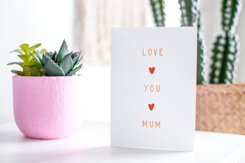 A greeting card featured standing up on a white tabletop with a pink plant pot in the background and some succulents in the pot. There’s a woven basket in the background with a cactus inside. The card features the words “Love You Mum” with a small, red heart in between each word. 