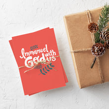 Load image into Gallery viewer, A stack of Christmas cards with brown string wrapped around them. A brown craft paper gift is off to the side. The card has a light red background with the words &quot;Immanuel God with Us&quot; in white with a couple of plant leaves in navy around the words.