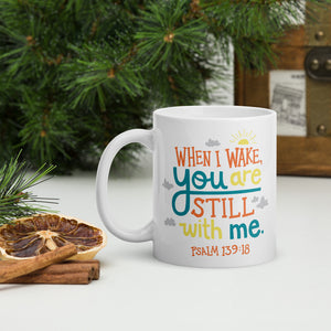 A white mug sits on a table, surrounded by evergreen foliage, dried lemon and cinnamon sticks. The mug reads 'When I wake you are still with me, Psalm 139:18' in orange, teal and yellow lettering, illustrated with a small yellow sun and little grey clouds. 