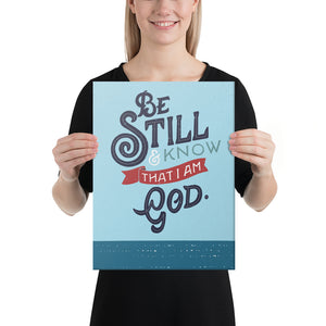 A woman is holding an art print in front of her and smiling. The print is bright blue with the verse 'Be Still and Know that I am God' illustrated in a bold typographic style. The woman is white with blond hair and wearing black.