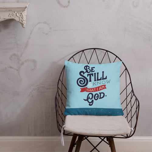 A blue cushion sits on a wire frame chair against a neutral plastered wall. The cushion is bright blue with the verse 'Be Still and Know that I am God' illustrated in a bold typographic style.