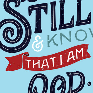 Closeup on a bright blue art print with black writing and red accent, featuring a section of the verse 'Be Still and know that I am God'