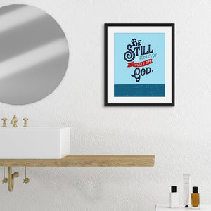 A framed art print hanging on a white wall in a calm bathroom scene. There's also a round mirror, wooden shelf, white sink, and some toiletries. The print is bright blue with the verse 'Be Still and Know that I am God' illustrated in a bold typographic style.