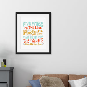 Framed artwork in a black frame on a wall above a sofa featuring a white paper print with colorful lettering with the Bible verse Psalm 105:1 "Give praise to the Lord, proclaim his name; make known among the nations what he has done."