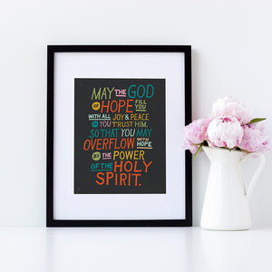 Artwork in a black frame with the with a white matte. The artwork is on a black background with colorful letters reading May the God of hope fill you with all joy and peace as you trust him, so that you may overflow with hope by the power of the holy spirit." 