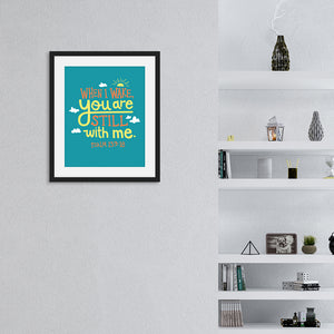 A turquoise print in a black frame hangs on a white living room wall. The print reads 'When I wake you are still with me, Psalm 139:18' in orange and yellow lettering, illustrated with a small yellow sun and little grey clouds. Beside the print is a set of shelves holding monochrome ornaments.