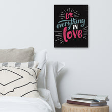 Load image into Gallery viewer, A black canvas hangs on a white bedroom wall. The canvas reads &quot;Do everything in love&quot; in bright pink and blue hand-lettering style, with white dashes around the words. Beneath it is a white bed covered with pillows, and a wooden sidetable with a pile of books.
