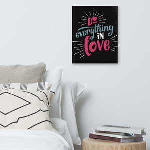 A black canvas hangs on a white bedroom wall. The canvas reads "Do everything in love" in bright pink and blue hand-lettering style, with white dashes around the words. Beneath it is a white bed covered with pillows, and a wooden sidetable with a pile of books.