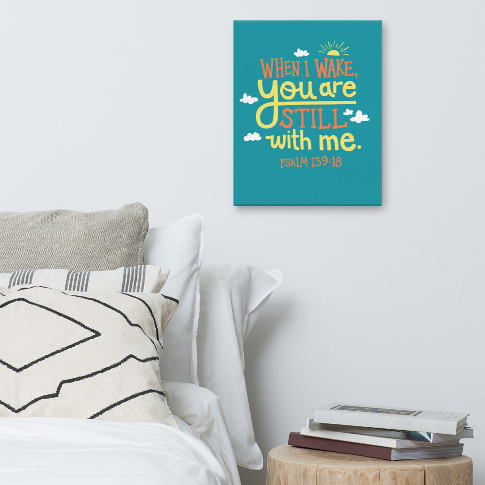 A turquoise canvas hangs on a white bedroom wall, above a white bed covered with gery and white pillows, and a small wooden table holding books. The canvas reads 'When I wake you are still with me, Psalm 139:18' in orange and yellow lettering, illustrated with a small yellow sun and little grey clouds.