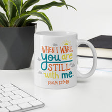 Load image into Gallery viewer, A white mug sits on a desk, surrounded by books, a pot plant and a white computer keyboard. The mug reads &#39;When I wake you are still with me, Psalm 139:18&#39; in orange, teal and yellow lettering, illustrated with a small yellow sun and little grey clouds. 