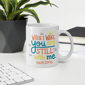 A white mug sits on a desk, surrounded by books, a pot plant and a white computer keyboard. The mug reads 'When I wake you are still with me, Psalm 139:18' in orange, teal and yellow lettering, illustrated with a small yellow sun and little grey clouds. 