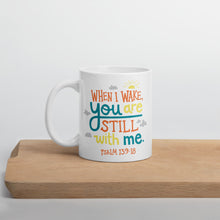 Load image into Gallery viewer, A white mug sits on a light wooden board, in front of a white wall. The mug reads &#39;When I wake you are still with me, Psalm 139:18&#39; in orange, teal and yellow lettering, illustrated with a small yellow sun and little grey clouds. 