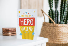 Load image into Gallery viewer, A greeting card is featured on a white tabletop with a white planter in the background with a green plant. There’s a woven basket in the background with a cactus inside. The card features illustrated lettering reading “You are my hero mum” with stars around it. There’s a background behind the word “mum” featuring yellow stripes and the word “hero” has a red background. 