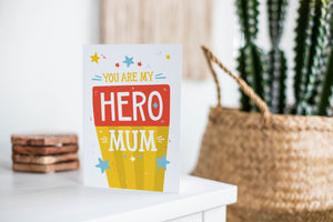 A greeting card is featured on a white tabletop with a white planter in the background with a green plant. There’s a woven basket in the background with a cactus inside. The card features illustrated lettering reading “You are my hero mum” with stars around it. There’s a background behind the word “mum” featuring yellow stripes and the word “hero” has a red background. 