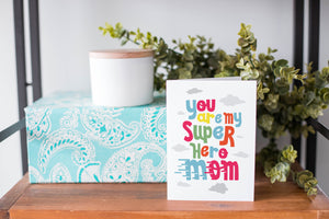 A greeting card is on a table top with a present in blue wrapping paper in the background. On top of the present is a candle and some greenery from a plant too. The card features the words “You are my super hero mom.”