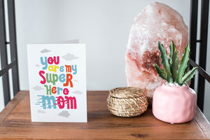 A card on a wood tabletop and on the right side of the card is a woven basket, a pink plant pot with a cactus in it and a pink crystal rock. The card features the words “You are my super hero mom.”