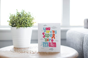 A greeting card is featured on a wood coffee table with a green plant in a white planter in the background. The card features the words “You are my super hero mum” with clouds in the background. 