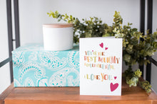 Load image into Gallery viewer, A greeting card is on a table top with a present in blue wrapping paper in the background. On top of the present is a candle and some greenery from a plant too. The card features illustrated lettering reading “You’re the best mummy I could ever have. I love you” with hearts surrounding the words. 