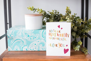 A greeting card is on a table top with a present in blue wrapping paper in the background. On top of the present is a candle and some greenery from a plant too. The card features illustrated lettering reading “You’re the best mummy I could ever have. I love you” with hearts surrounding the words. 