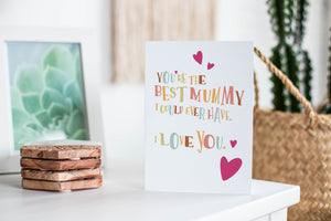 A greeting card featured standing up on a white tabletop with a framed photo of a succulent in the background and a stack of wooden coasters. There’s a woven basket in the background with a cactus inside. The card features illustrated lettering reading “You’re the best mummy I could ever have. I love you” with hearts surrounding the words. 
