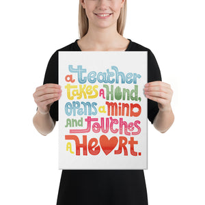 A woman holding a canvas in her hands with the words “A teacher takes a hand, opens a mind, and touches a heart” on the canvas. The “a” in the word “heart” is a heart shape and the words are blue, red, yellow and green. 