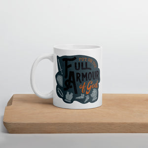 A white mug sits on a wooden board against a white backdrop. The mug features the quote 'Put on the full armour of God' in black and orange typography, along with illustrated pieces of armour in medieval style against a dark gray background.