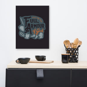 A dark gray canvas hangs on a white kitchen wall, over black cups and wooden utensils sitting on a black sideboard. The canvas features the quote 'Put on the full armour of God' in black and orange typography, with medieval-style pieces of armour illustrated in pale gray.