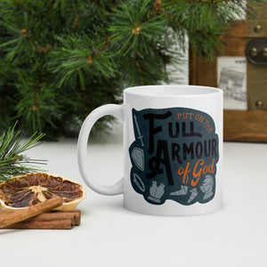 A white mug sits on a white table surrounded by evergreen branches, dried oranges and cinnamon. The mug features the quote 'Put on the full armour of God' in black and orange typography, along with illustrated pieces of armour in medieval style against a dark gray background.