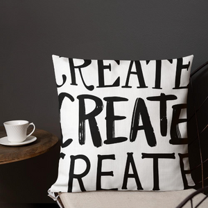 A pillow on a chair with a coffee mug on a table next to it. The white pillow features the phrase “create, create, create" in black lettering with each word in three different fonts. 