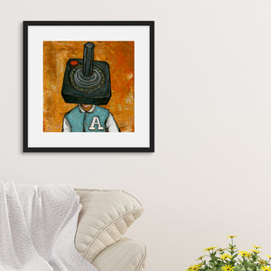 A black frame on a wall featuring an illustration of an Atari controller. The frame is above the corner of a sofa.