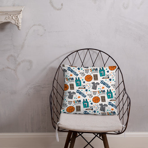 A pillow on a chair against a grey wall. The pillow is white features hand drawn illustrations of basketball themed items.