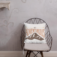 Load image into Gallery viewer, A pillow on a chair in front of a wall. The white pillow features the phrase “Be adventurous” with arrows pointing to the word “be” and a brown mountain illustration underneath the word “adventure.”