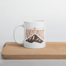 Load image into Gallery viewer, A white mug sitting on a piece of wood. The quote on the mug says “Be adventurous” with arrows pointing to the word “be” and a brown mountain illustration underneath the word “adventure.”