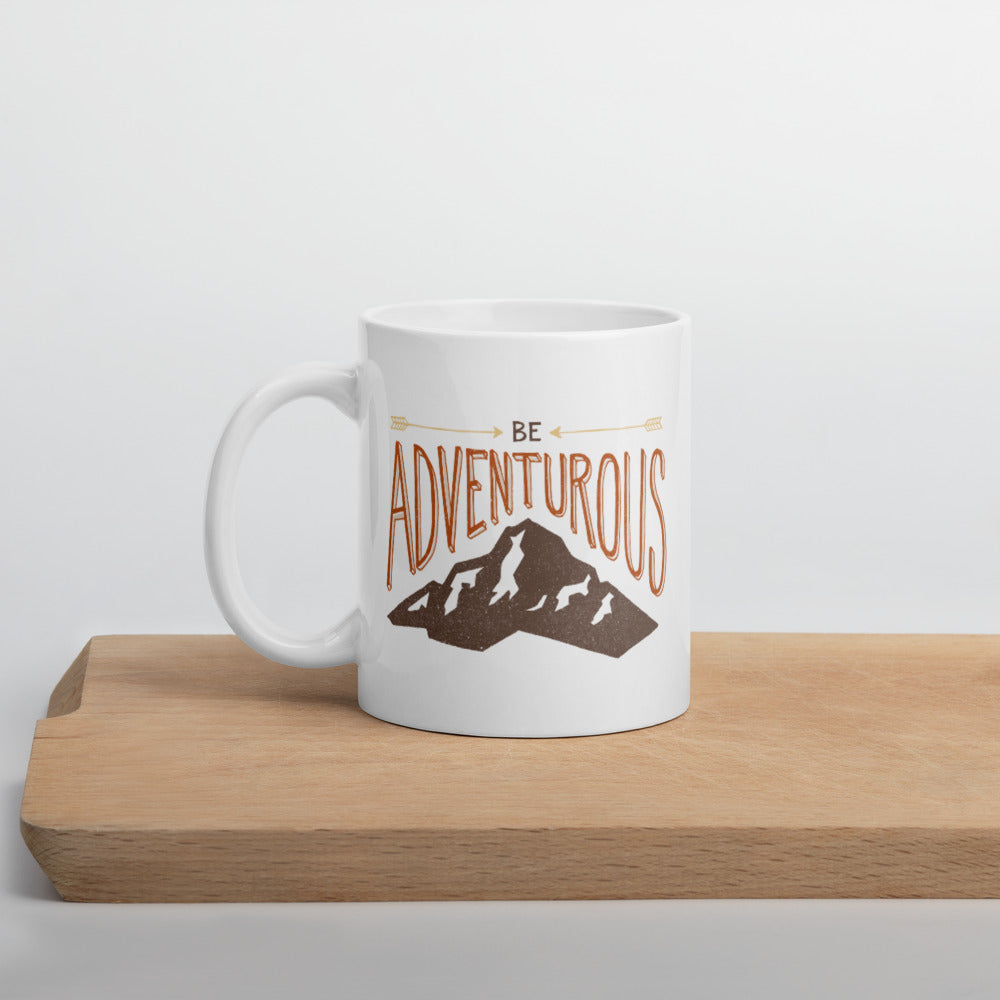 A white mug sitting on a piece of wood. The quote on the mug says “Be adventurous” with arrows pointing to the word “be” and a brown mountain illustration underneath the word “adventure.”