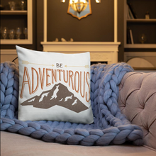Load image into Gallery viewer, A pillow on a sofa with a blue knitted blanket with the phrase “Be adventurous” with arrows pointing to the word “be” and a brown mountain illustration underneath the word “adventure.” The pillow is white. 