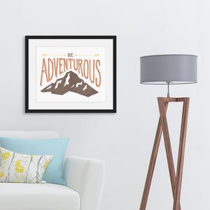 A black frame hanging on the wall above a sofa. The lettering and illustration says “Be adventurous” with arrows pointing to the word “be” and a brown mountain illustration underneath the word “adventure.”